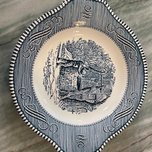 May include: A ceramic bowl with a blue and white transferware pattern. The center features a detailed illustration of a person near a well and a house. The wide rim is decorated with blue scrollwork and a repeating dotted border design.
