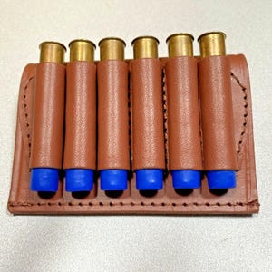 May include: A brown leather shotgun shell holder with six brass and blue shells. The holder is rectangular with stitched edges, designed to carry ammunition. The shells are arranged vertically, showcasing the product's functionality and design.