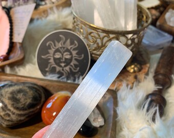 May include: A white selenite wand, also known as a cleansing wand, is held in a hand. The wand is smooth and has a striated texture. It is a popular crystal for energy clearing and meditation.