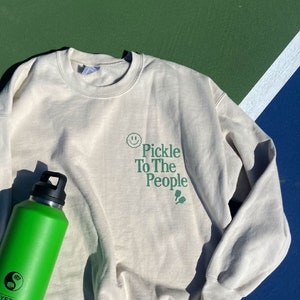 Puede incluir: Una sudadera beige claro con el texto "Pickle To The People" en letras verdes. Una botella de agua Yeti verde con un s&iacute;mbolo yin yang est&aacute; en el lado izquierdo de la sudadera.