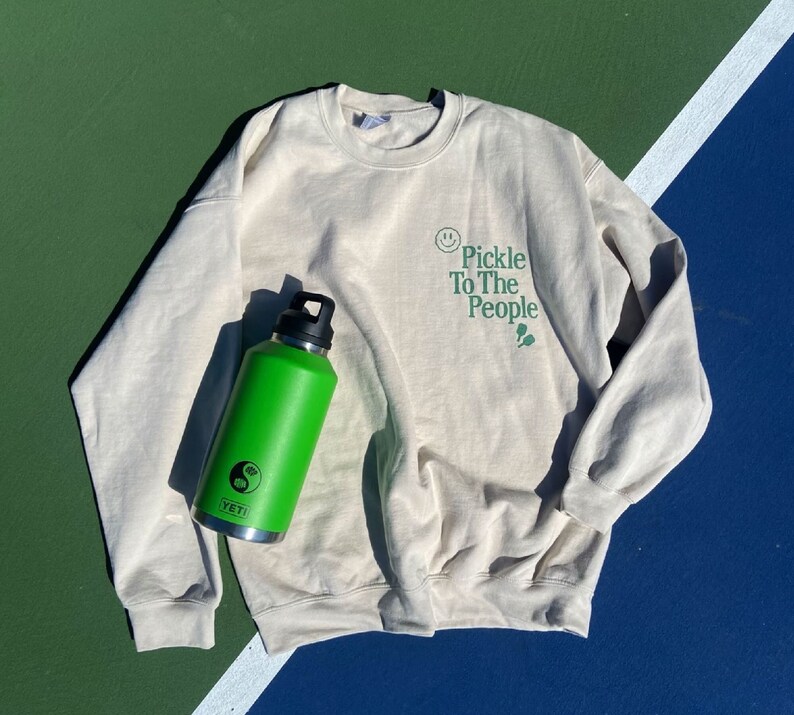Puede incluir: Una sudadera beige claro con el texto "Pickle To The People" en letras verdes. Una botella de agua Yeti verde con un s&iacute;mbolo yin yang est&aacute; en el lado izquierdo de la sudadera.