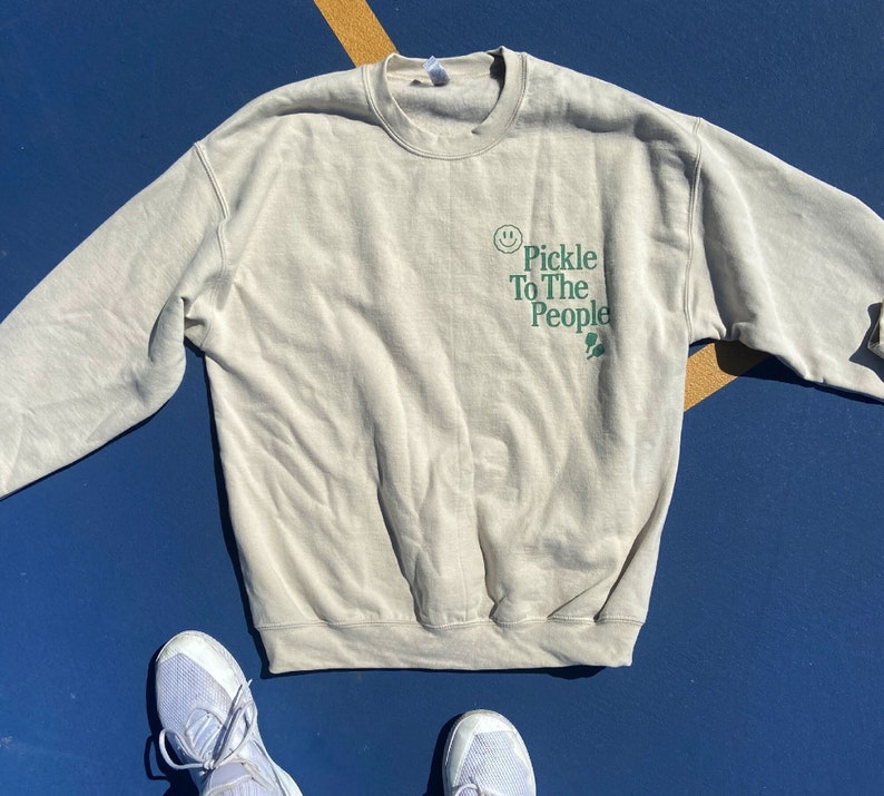 Puede incluir: Una sudadera beige claro con un gr&aacute;fico verde que dice "Pickle To The People" con una carita sonriente y una hoja verde.
