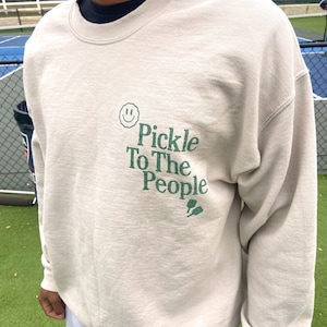 Puede incluir: Una sudadera de color beige claro con una carita sonriente verde y el texto "Pickle To The People" en verde. La sudadera tiene cuello redondo y mangas largas.