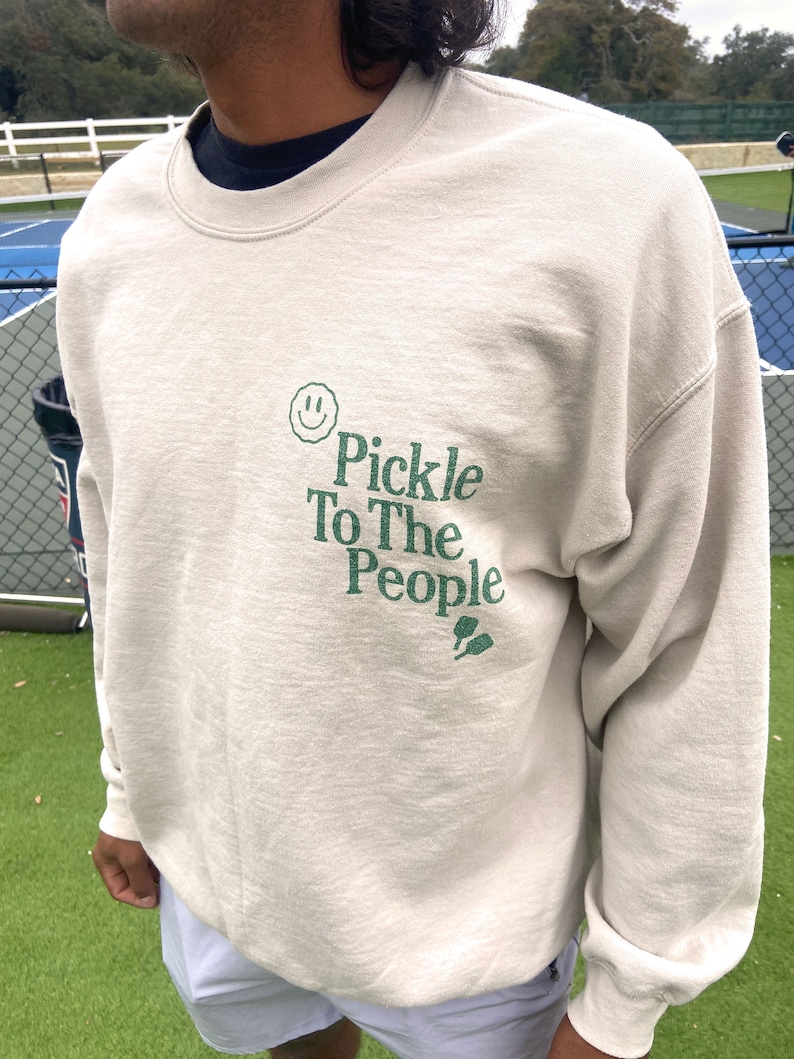 Puede incluir: Una sudadera de color beige claro con una carita sonriente verde y el texto "Pickle To The People" en verde. La sudadera tiene cuello redondo y mangas largas.