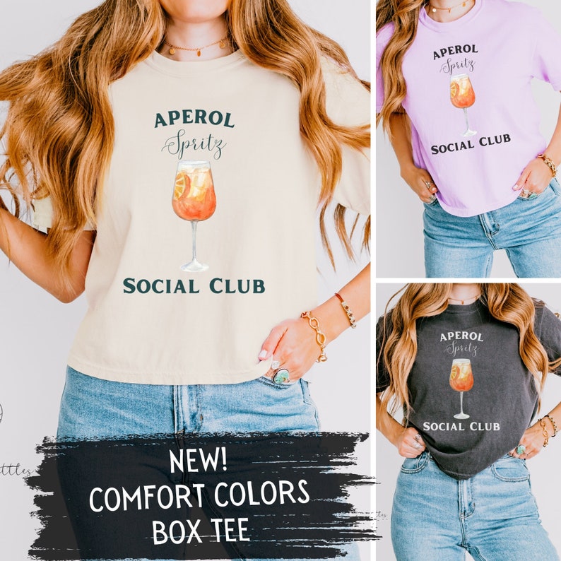 May include: Comfort Colors box tee in cream, lilac, and charcoal gray. Each shirt features a watercolor illustration of an Aperol Spritz cocktail with the text "APEROL Spritz SOCIAL CLUB". The bottom of the image reads "NEW! COMFORT COLORS BOX TEE".