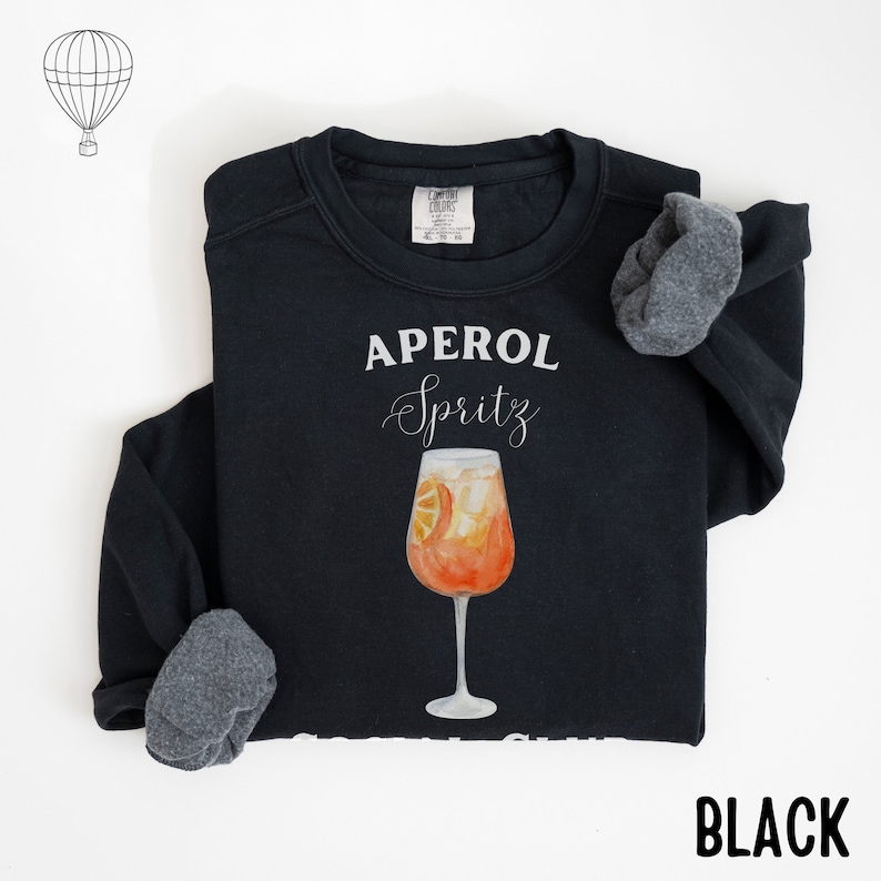 May include: Black sweatshirt with the words "APEROL Spritz" in white script above a watercolor illustration of the cocktail. The image includes an orange slice and ice in a stemmed glass. The word "BLACK" is printed at the bottom.