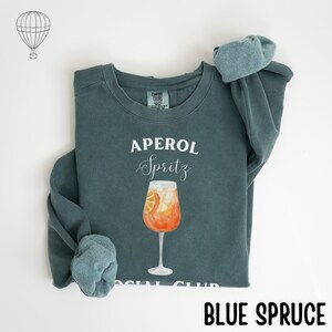 May include: Blue spruce sweatshirt with the words "APEROL Spritz" in white script above a watercolor illustration of an Aperol Spritz cocktail in a tall glass. The words "SOCIAL CLUB" are at the bottom.