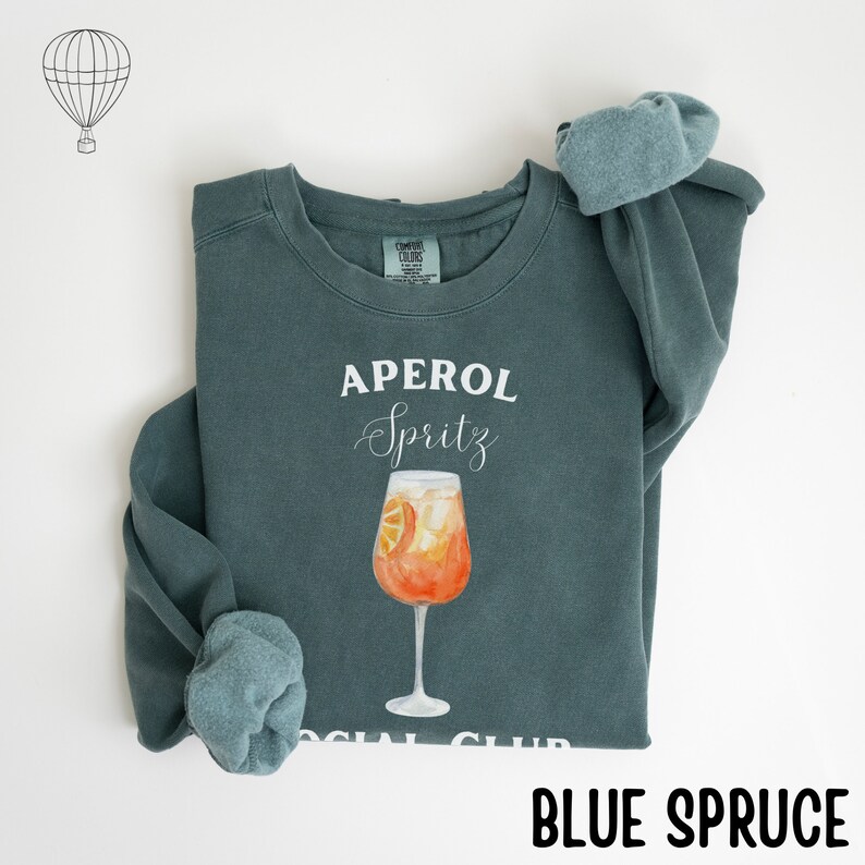 May include: Blue spruce sweatshirt with the words "APEROL Spritz" in white script above a watercolor illustration of an Aperol Spritz cocktail in a tall glass. The words "SOCIAL CLUB" are at the bottom.