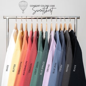 May include: A rack displays a variety of Comfort Colors 1566 sweatshirts in various colors, including white, butter, terracotta, watermelon, crimson, light green, blue spruce, orchid, blue jean, denim, pepper, and black. The word "Sweatshirt" is written in cursive.