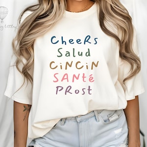 May include: Cream-colored t-shirt with the words "Cheers, Salud, Cin Cin, Santé, Prost" in various colors. The shirt has a crew neck and short sleeves. The text is in a playful, handwritten style.