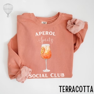 May include: Terracotta-colored sweatshirt with the words "APEROL Spritz SOCIAL CLUB" in white. A watercolor illustration of an Aperol Spritz cocktail is centered on the front. A matching scrunchie is next to the sweatshirt. The sweatshirt is made of a soft material.
