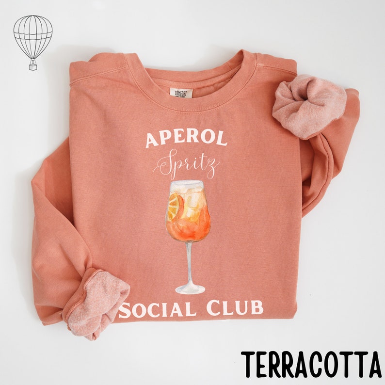 May include: Terracotta-colored sweatshirt with the words "APEROL Spritz SOCIAL CLUB" in white. A watercolor illustration of an Aperol Spritz cocktail is centered on the front. A matching scrunchie is next to the sweatshirt. The sweatshirt is made of a soft material.
