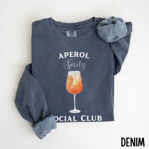 May include: A denim blue sweatshirt with the words "APEROL Spritz SOCIAL CLUB" in white. A watercolor illustration of an Aperol Spritz cocktail in a stemmed glass is centered on the front. A matching scrunchie rests on the shoulder.