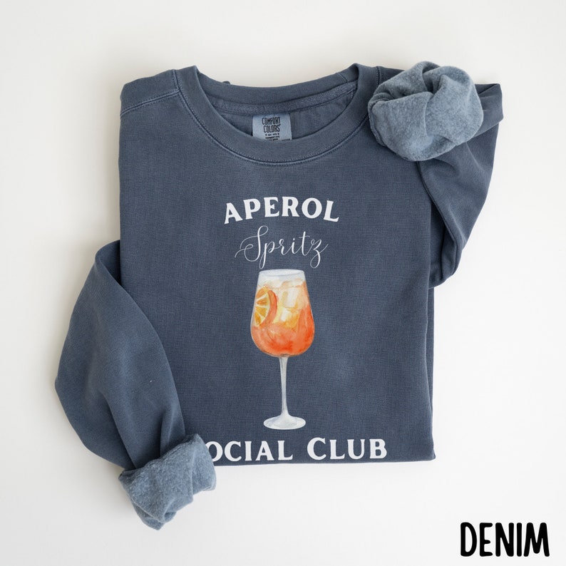 May include: A denim blue sweatshirt with the words "APEROL Spritz SOCIAL CLUB" in white. A watercolor illustration of an Aperol Spritz cocktail in a stemmed glass is centered on the front. A matching scrunchie rests on the shoulder.