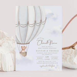 Peut inclure: Invitation de baby shower avec un ballon à air chaud et un ours en peluche. Le texte "We're on Cloud Nine" est présent, ainsi que les détails de la baby shower d'Hannah & Thomas, incluant la date, l'heure et le lieu.