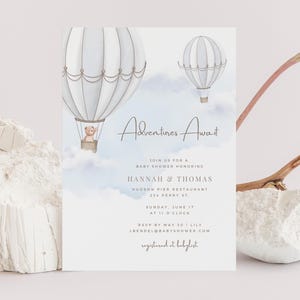 May include: A baby shower invitation with a watercolor design. Two hot air balloons float in a blue sky with the words "Adventures Await." The invitation includes details for Hannah & Thomas's baby shower, including the date, time, and location.