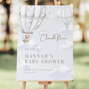 Peut inclure: Panneau de bienvenue pour une baby shower sur un chevalet. Le panneau présente deux montgolfières, dont une avec un ours en peluche dans le panier. Le texte comprend "We're on Cloud Nine" et "Hannah's Baby Shower". Le fond est bleu clair avec des nuages.