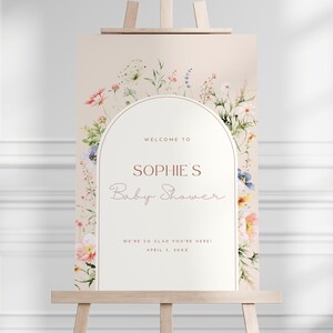 May include: A welcome sign for a baby shower with a floral arch design. The sign is white with a peach background and says "Welcome to Sophie's Baby Shower. We're so glad you're here! April 2, 20XX"