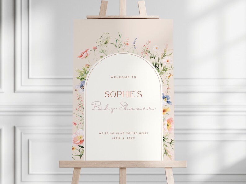 May include: A welcome sign for a baby shower with a floral arch design. The sign is white with a peach background and says "Welcome to Sophie's Baby Shower. We're so glad you're here! April 2, 20XX"