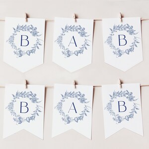 May include: A white banner with blue floral wreaths and the letters "BAB" in the centre of each wreath. The banner is strung with twine and clothes pegs.