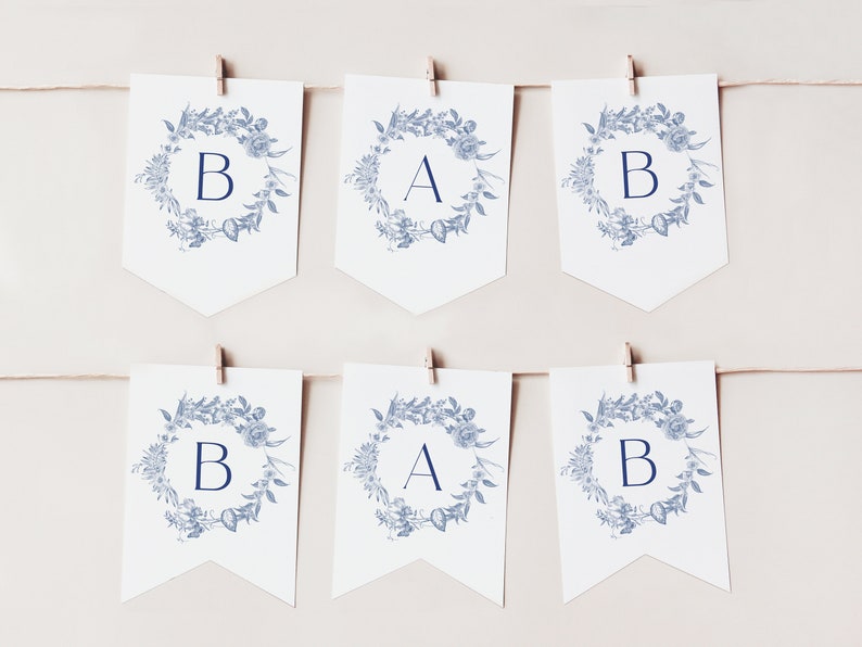 May include: A white banner with blue floral wreaths and the letters "BAB" in the centre of each wreath. The banner is strung with twine and clothes pegs.