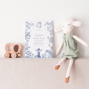 May include: A white invitation card with blue floral designs and a giraffe illustration. The card says "Join us for a baby shower" and "Daisy Avery". A wooden toy elephant and a white stuffed lamb are sitting on a beige couch.
