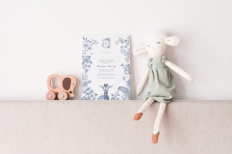 May include: A white invitation card with blue floral designs and a giraffe illustration. The card says "Join us for a baby shower" and "Daisy Avery". A wooden toy elephant and a white stuffed lamb are sitting on a beige couch.