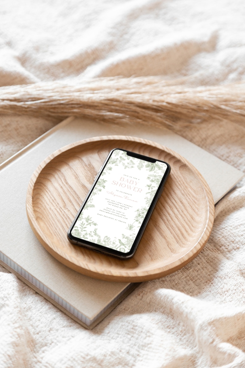 May include: A smartphone displaying a baby shower invitation with a floral design. The invitation reads "Join us for a Baby Shower in celebration of [Name]" and includes the date, time, and location of the event.