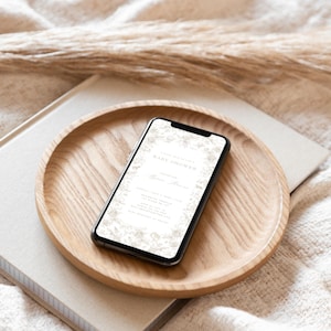 May include: A smartphone displaying a baby shower invitation with a floral design. The invitation includes the text "Celebrating the Baby Shower" and the name "Emilia". The phone is resting on a wooden tray.
