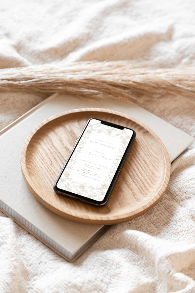 May include: A smartphone displaying a baby shower invitation with a floral design. The invitation includes the text "Celebrating the Baby Shower" and the name "Emilia". The phone is resting on a wooden tray.