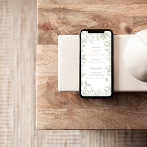 May include: A smartphone displaying a baby shower invitation with a floral design. The invitation reads "Join us for a Baby Shower" and includes the name of the person being celebrated, the date, time, and location of the event.