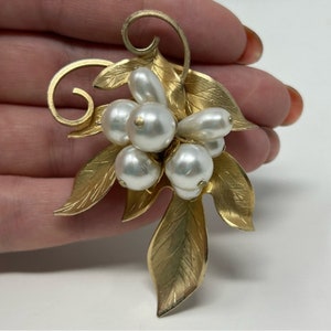 Puede incluir: Un broche dorado con un racimo de perlas falsas sobre un diseño de hoja.