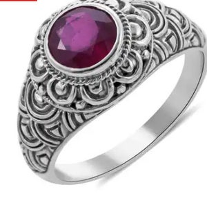 May include: Sterling silver ring featuring a large, round, deep red ruby gemstone. The ring has an ornate, detailed design around the gemstone and along the band. The intricate patterns and the vibrant color of the ruby create a striking contrast.