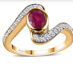 May include: A gold-toned ring with a large oval ruby center stone. The ring features a wave-like design, with a band of small, clear stones accenting the curves. The inscription "D'joy 925" is visible on the band.