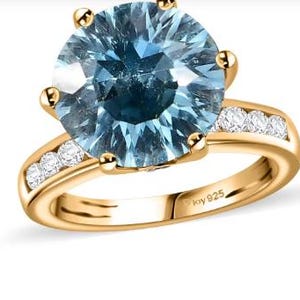 Może przedstawiać: Złoty pierścionek z dużym niebieskim kamieniem szlachetnym i mniejszymi, przezroczystymi kamieniami szlachetnymi osadzonymi w obręczy wokół pierścionka. Pierścionek jest oznaczony "925" od wewnątrz.