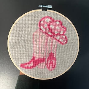 Könnte beinhalten: Rosa Cowboy-Stiefel und ein rosa-weiß gepunkteter Hut, gestickt auf einem beigefarbenen Stoff in einem hölzernen Stickrahmen.