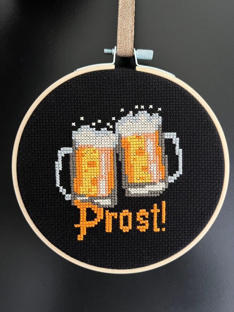 May include: A black embroidery hoop with a cross-stitch design of two beer steins clinking together with the word "Prost!" in orange thread.