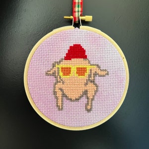 Peut inclure: Un cercle de broderie rond en point de croix, de couleur violette et blanche, avec une dinde portant un chapeau rouge et des lunettes de soleil. La dinde est brune et a un corps blanc. Le cercle est suspendu à un ruban rouge et blanc à carreaux.