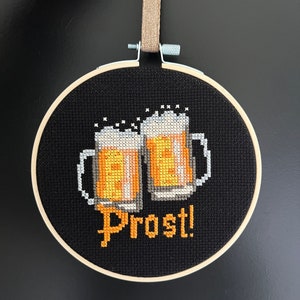 May include: A black embroidery hoop with a cross-stitch design of two beer steins and the word "Prost!" in orange thread.