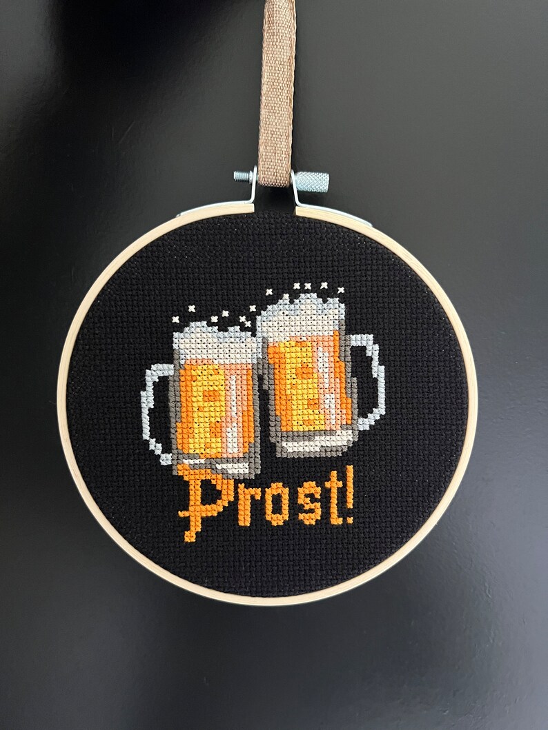 May include: A black embroidery hoop with a cross-stitch design of two beer steins and the word "Prost!" in orange thread.