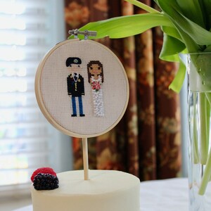 Peut inclure: Un gâteau blanc avec une seule fleur blanche sur le dessus. La fleur est fixée à un bâton en bois et a un petit cerceau brodé avec un couple en uniforme militaire et une robe de mariée. Le gâteau est décoré d'une seule mûre.
