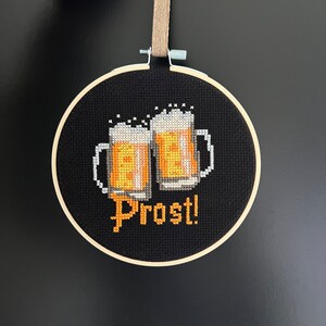 May include: A black embroidery hoop with a cross-stitch design of two beer steins and the word "Prost!" in orange thread.