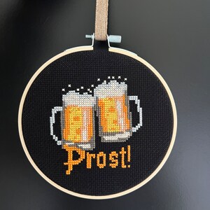 May include: A black embroidery hoop with a cross-stitch design of two beer steins and the word "Prost!" in yellow thread.