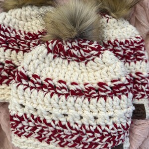 May include: Three cream-colored crocheted beanies with alternating red stripes and fluffy brown pom-poms. Each hat has a small brown leather tag. The hats are displayed on a pink surface.