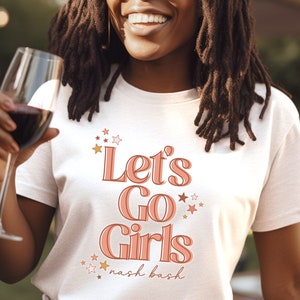 May include: White t-shirt with the text "Let's Go Girls" in pink and orange lettering with stars. The text "nash bash" is written in smaller letters below the main text.