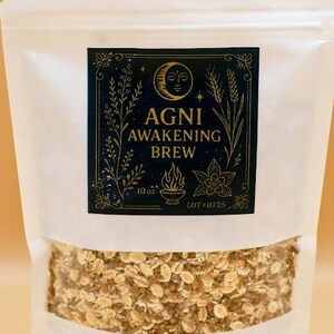 May include: A white pouch filled with a granular product, labeled "AGNI AWAKENING BREW." The label is black with gold accents, featuring a crescent moon, floral elements, and the text "10 oz." The pouch is set against a light brown background.