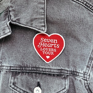 May include: A red heart-shaped patch with white embroidered text reading "Seven Hearts LOVERS TOUR" on a gray denim jacket. The patch has a white border and a small white heart at the bottom.