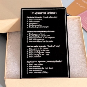 May include: A black rectangular card featuring white text titled The Mysteries of the Rosary, listing the Joyful, Luminous, Sorrowful, and Glorious mysteries with their respective days. The card rests inside a light brown cardboard box with white foam.
