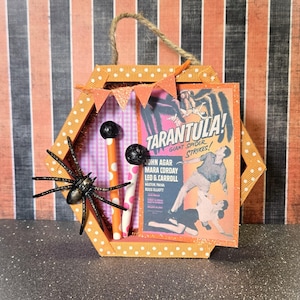 May include: A Halloween-themed, orange and white polka-dotted, hexagonal frame with a decorative spider and a vintage movie poster that reads "Tarantula! Giant Spider Strikes!". The frame also features two decorative items and small orange pennants.