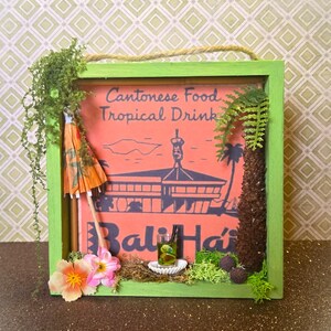 May include: A framed sign with the text "Cantonese Food Tropical Drink" and "Bali Hai" in a retro style. The frame is green, with an orange background. An orange cocktail umbrella and miniature drink are included.